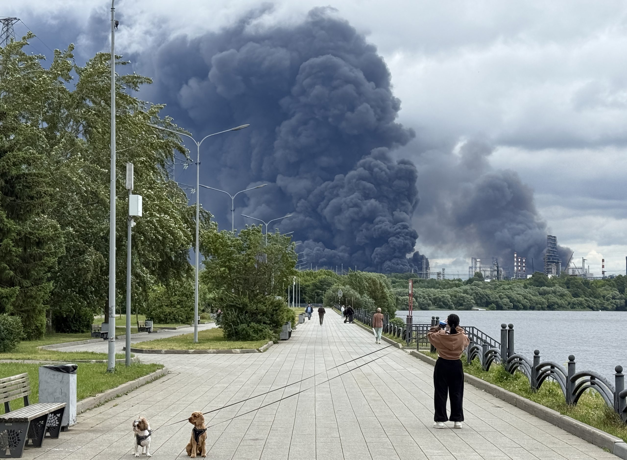 People walk in a park as black smoke rises from the area of the Russian oil producer Gazprom Neft's Moscow oil refinery on the south-eastern outskirts of Moscow on June 18, 2026. Moscow was fending off a "large-scale" drone attack from Ukraine, with several drones reaching an oil refinery, the city's mayor Sergey Sobyanin said early on June 18, 2026. (Photo by AFP)